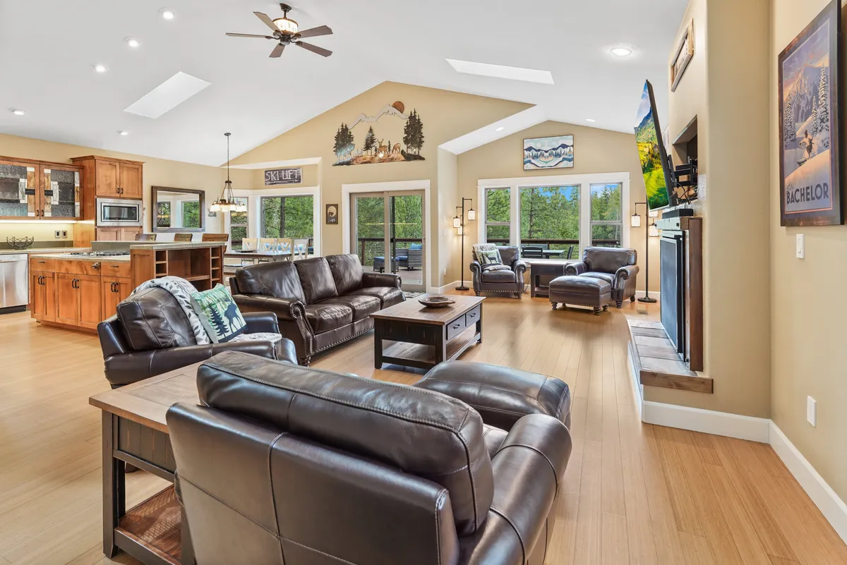 This image showcases a bright, open-concept living room furnished with comfortable leather sofas, large windows that offer outdoor views, wooden kitchen cabinetry, and a decorative mountain mural. The arrangement invites gathering, relaxation, and socializing, with clear sightlines between the living and dining areas.