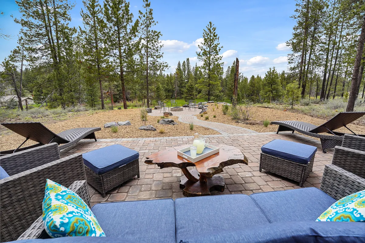 An inviting exterior lounge area with comfortable seating and a coffee table is nestled beneath the trees. The pathway in the background and natural surroundings suggest a seamless transition from structured spaces to the wild outdoors, while the tranquil mood encourages relaxation amidst nature. The area looks secure and friendly for pets to relax with their owners after a walk.