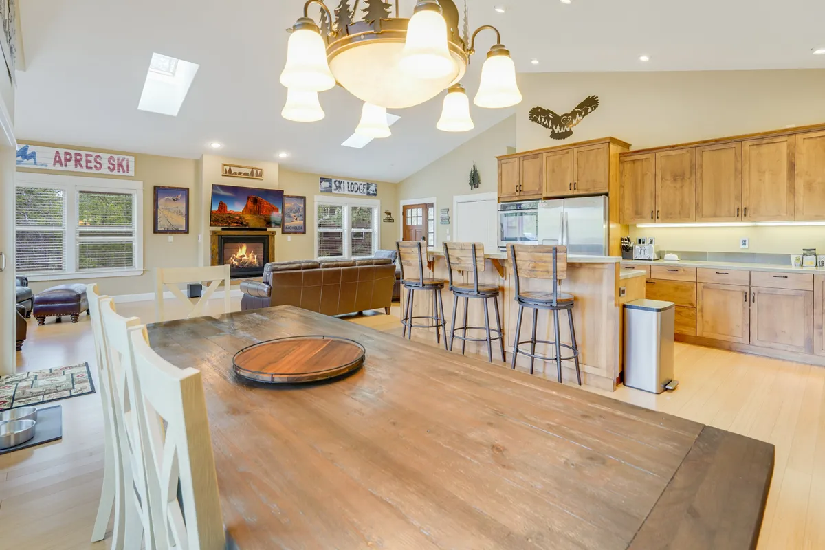 The image shows a large rustic wood dining table, adjacent kitchen island with bar stools, and a nearby fireplace. The dining area is open and welcoming, ideal for shared meals, board games, and gathering with loved ones.