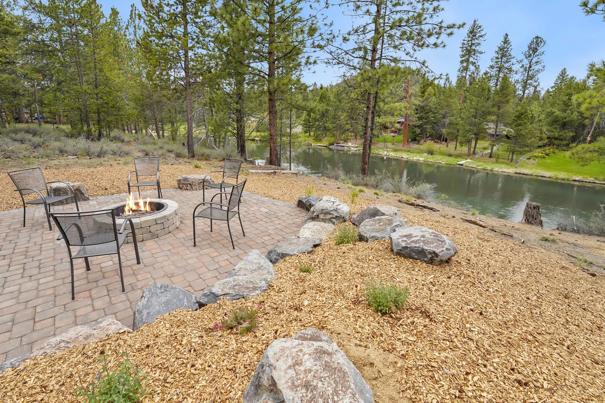 A cozy fire pit area with comfortable seating overlooks a serene waterfront, surrounded by lush natural landscape. The view highlights the close connection to the water and the forest beyond, creating an ideal spot for campfire gatherings after a day of river adventures.