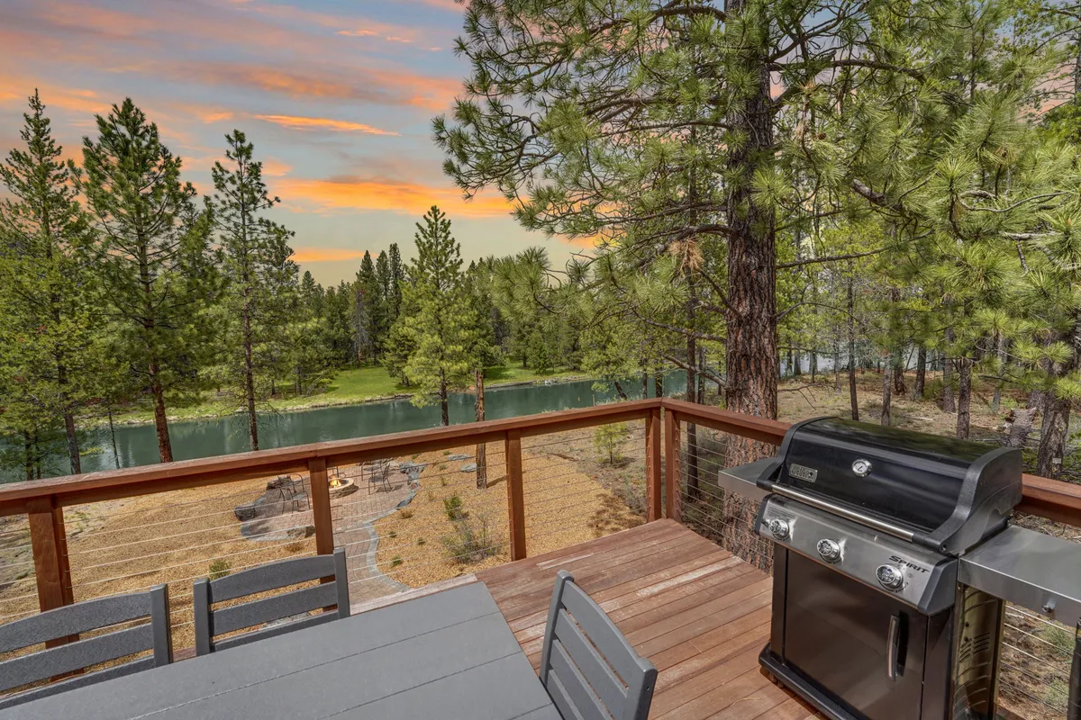 The image features an elevated deck overlooking the river at sunset, complete with a dining table, gas BBQ grill, and comfortable outdoor seating. The scene blends indoor comforts with breathtaking river views and hints at memorable meals shared outdoors.
