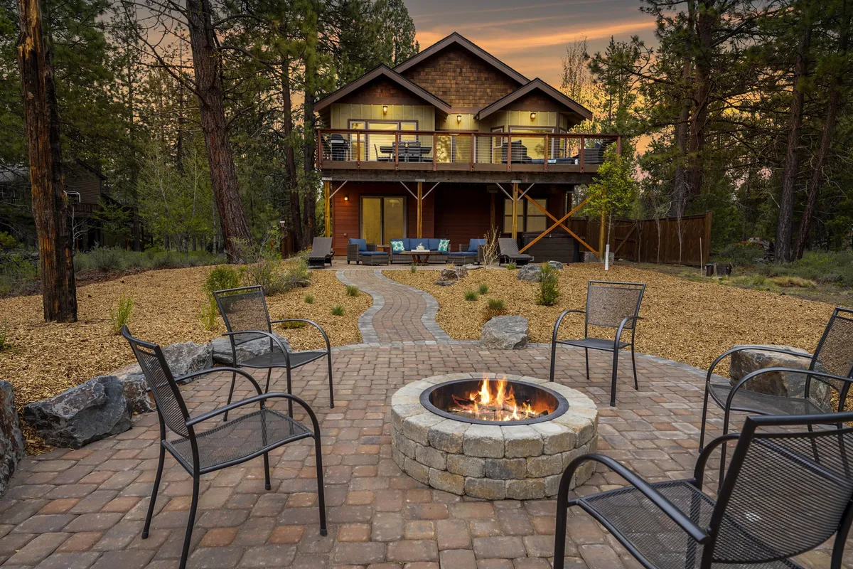 The image features the exterior of a charming single-family house nestled in a wooded setting along the Big Deschutes River. The focal points include a cozy fire pit surrounded by comfortable outdoor seating, a pathway leading up to the home, and inviting spaces perfect for gathering. The mood is serene and relaxed, with hints of outdoor dining and socializing under the open sky.