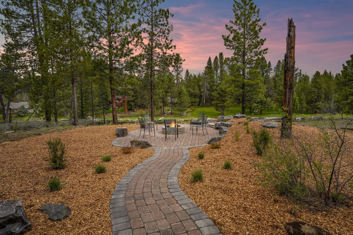 A cozy fire pit surrounded by curved seating and lush trees creates a welcoming evening gathering spot. The nearby pathway and abundant greenery suggest space for pets to roam, with the fire pit area offering a communal place for owners to enjoy evenings together. The mood is relaxed and cozy, ideal for winding down after a day of outdoor adventure.