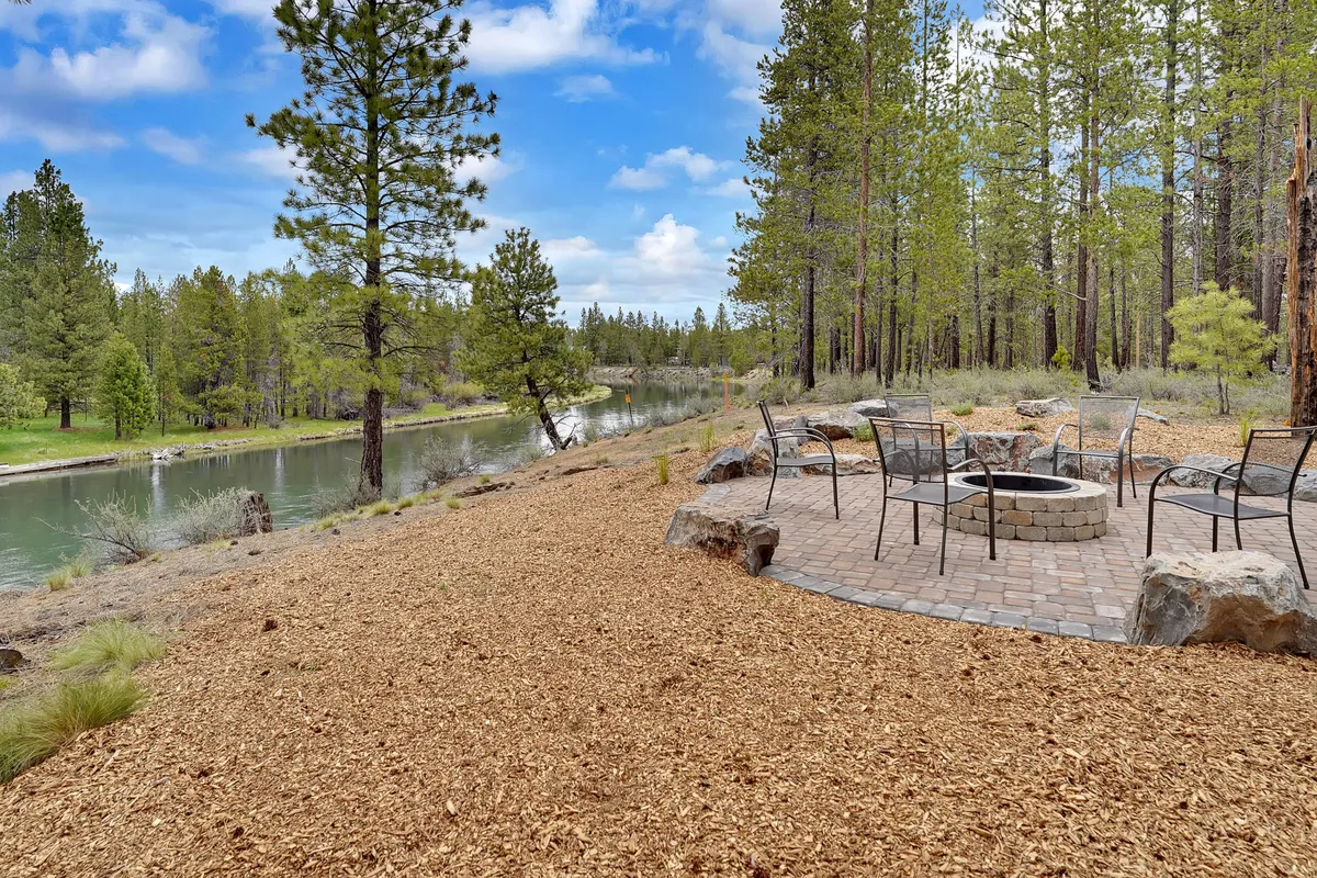 This exterior image features a fire pit set on the banks of a tranquil river, with mature trees providing shade and a sense of peaceful seclusion. The soft glow of the fire pit complements the natural setting, and the river's presence directly evokes the lodge's riverside location and the potential for fishing, kayaking, or tubing just steps away.