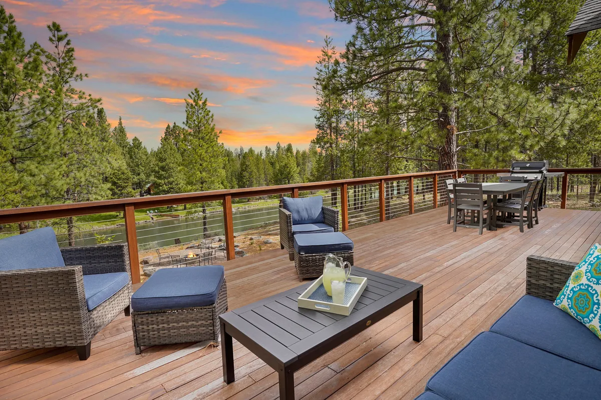 An elevated deck features a spacious outdoor dining table and comfortable seating, surrounded by forest views and a dramatic sunset sky. The area is perfect for summer gatherings, evening barbecues, and soaking in the scenery after a day of adventure.