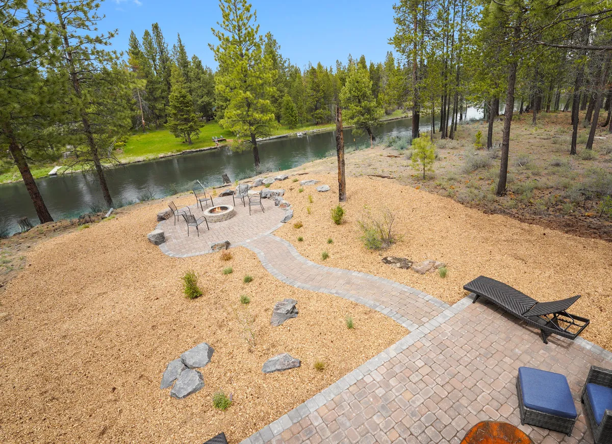 The image captures a riverfront fire pit area with ample seating, a tranquil river view, and a meandering pathway leading towards the water. The setting sun paints the sky, and the arrangement invites gatherings, s’mores, and nature appreciation.