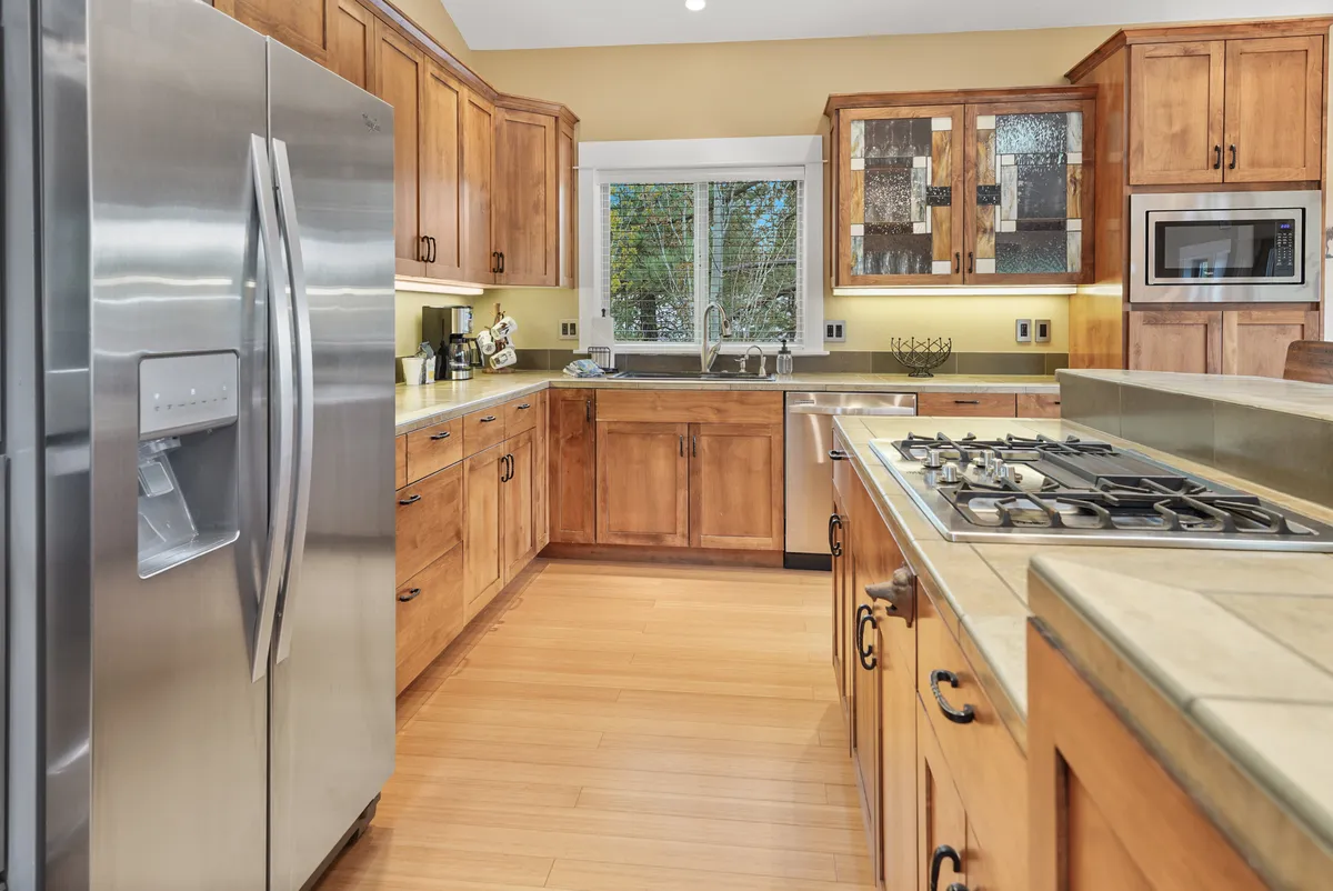 This image highlights a spacious, well-equipped kitchen with stainless steel refrigerator, kitchen island with stove, wooden cabinets, and a large window letting in natural light. The kitchen exudes warmth and functionality, perfect for both everyday meals and special gatherings.