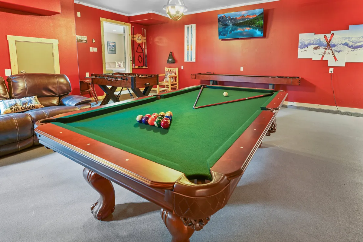 A multifunctional entertainment space with a pool table, leather couch, foosball table, large TV, and decorative mountain artwork. The room looks playful yet cozy, inviting both relaxation and activity.