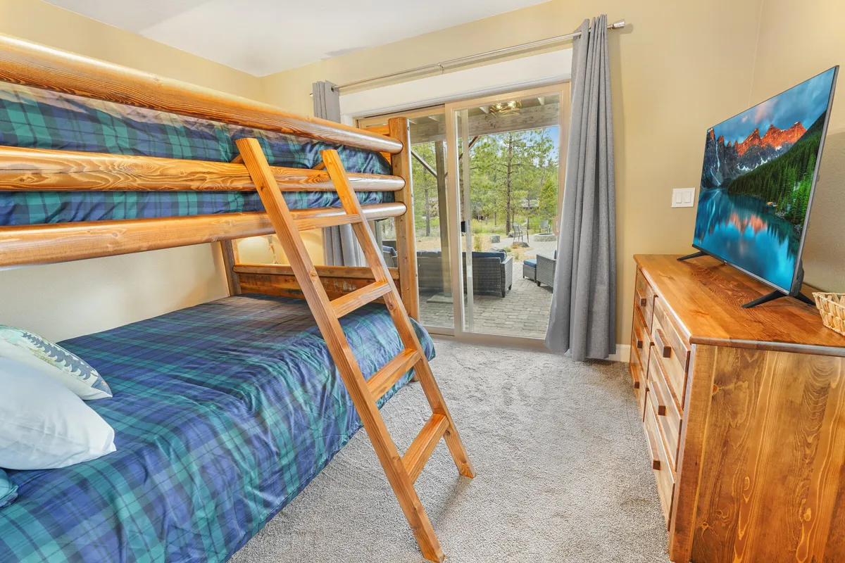 A queen-over-queen bunk bed with cozy bedding stands beside a dresser and a mounted television. The room features a sliding glass door that leads out to a patio area, perfect for easy access to outdoor relaxation.